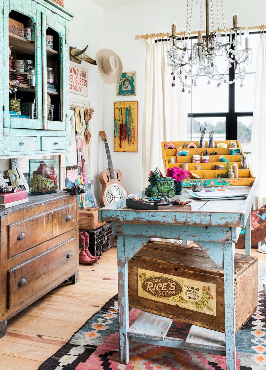 Espacio decorado con la tendencia cluttercore o desorden intencional. Hay una mesa central rústica pintada de color celeste y sobre ella distintos utensilios de arte. Al costado hay una vitrina con frascos y al fondo, un rincón de arte y música.