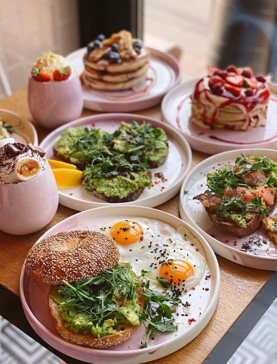 Brunch mostrando el detalle de una mesa de madera con vajilla rosada pastel. Sobre los platos hay todo para un brunch ideal: huevos, panes, panqueques y fruta.