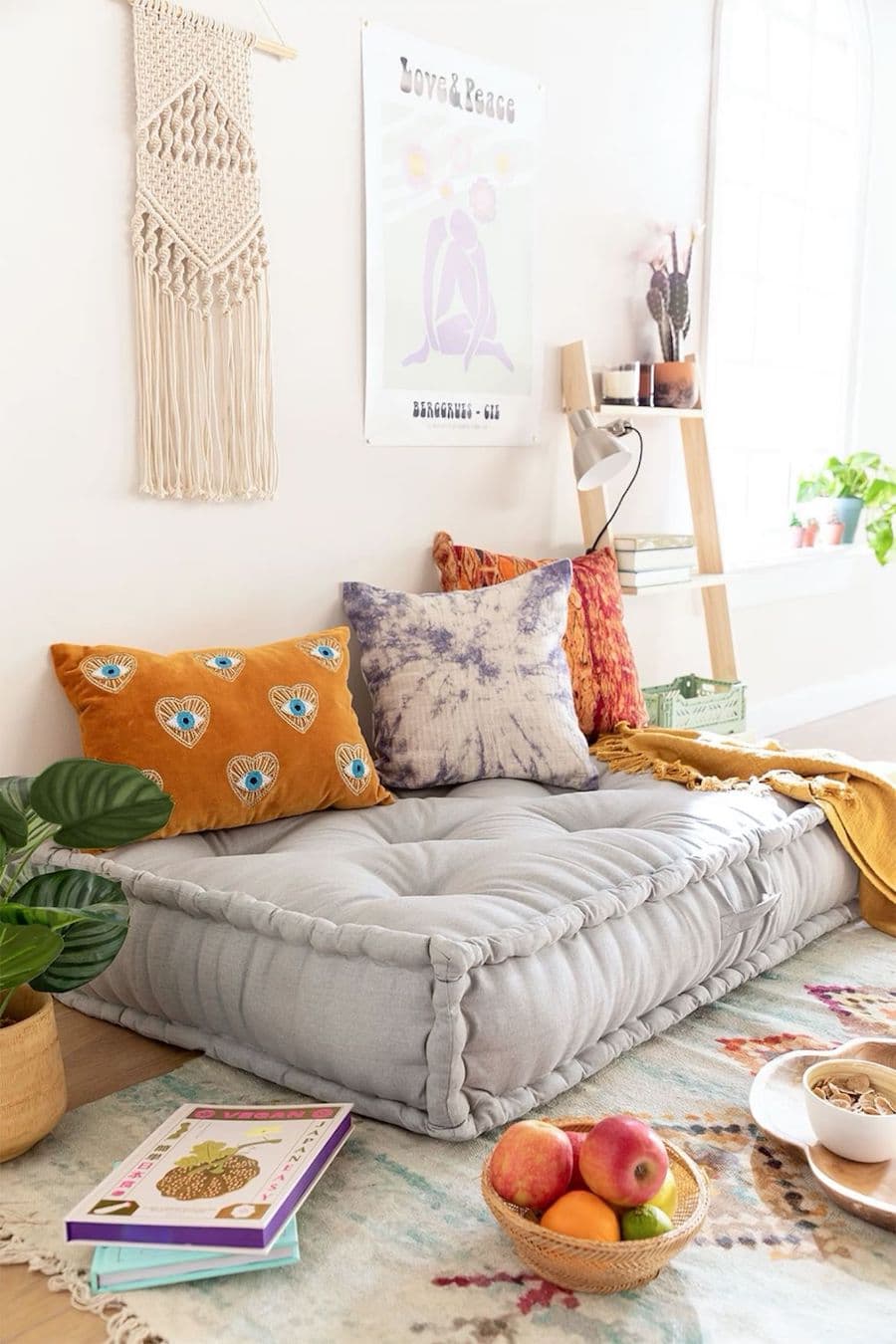 Rincón zen con gran cojín sobre el suelo. Sobre él hay cojines más pequeños de colores mostaza, blanco y gris. Los muros son claros y tiene mucha iluminación natural. 