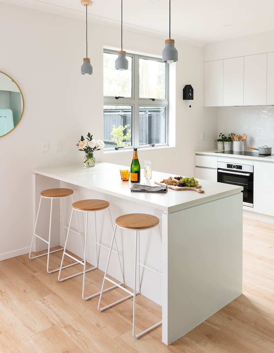 Cocina de muros y muebles blancos con una gran isla de cocina con 3 pisos de bar. Sobre la isla hay una tabla de picoteo, un espumante y copas. 