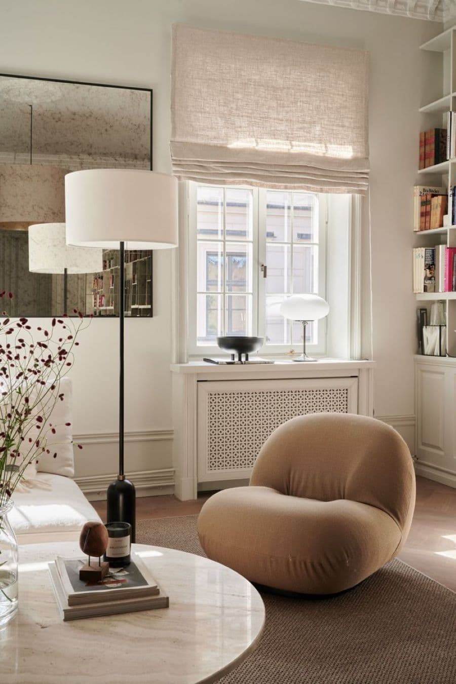 Living moderno con un sofá de líneas curvas, mullido de color beige oscuro. Ventana con cortina store de lino beige. Lámpara de pie con base negra y pantalla de tela blanca. En el muro hay un espejo grande envejecido.