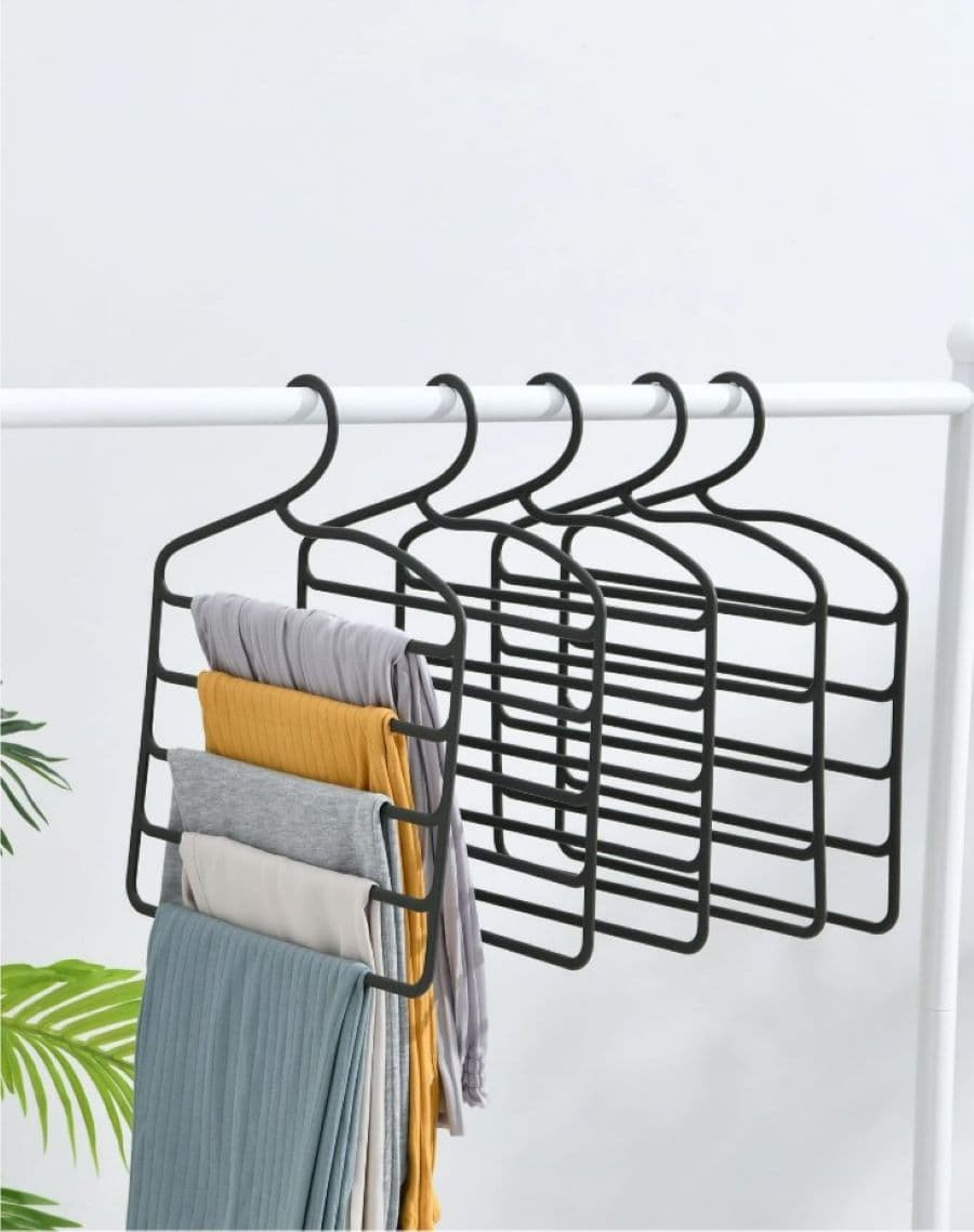 Colgador para pantalones con varios ganchos. 5 ganchos negros colgados en una barra blanca. Uno d ellos tiene 5 pantalones colgados, 3 jeans, uno de color beige y otro color mostaza. En la esquina se ve la rama de una planta