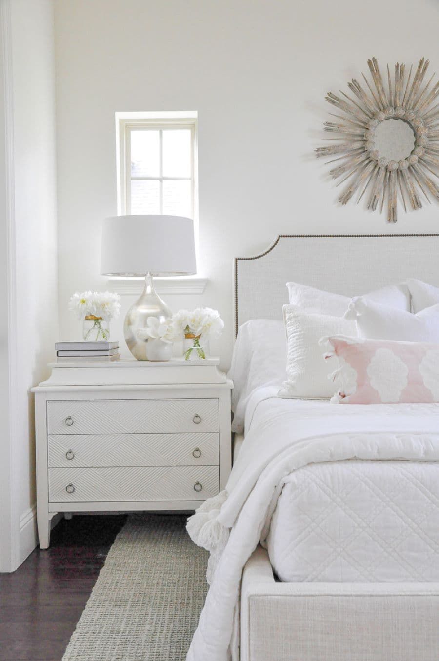 Dormitorio de estilo clásico. Gran cama con textiles blancos, sobre ella un espejo redondo color plateado. Velados de tres cajones con lámpara de base plateada, revistas y flores.