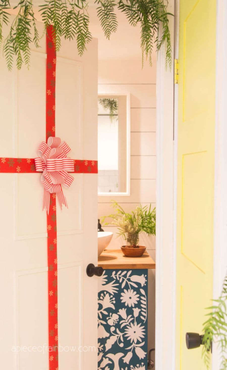 Puerta blanca con una cinta roja alrededor y un moño al centro. En la parte interior se ve un mueble con dibujos de flores azul y blanco. En la parte superior se ven ramas verdes colgando