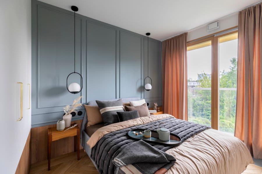 Ventana con cortinas de tela color durazno. Dormitorio con decoración en tonos grises y blancos, con cama vestida con colchas grises, almohadas y cojines coordinados, un banco de madera a los pies de la cama, y una lámpara colgante blanca.