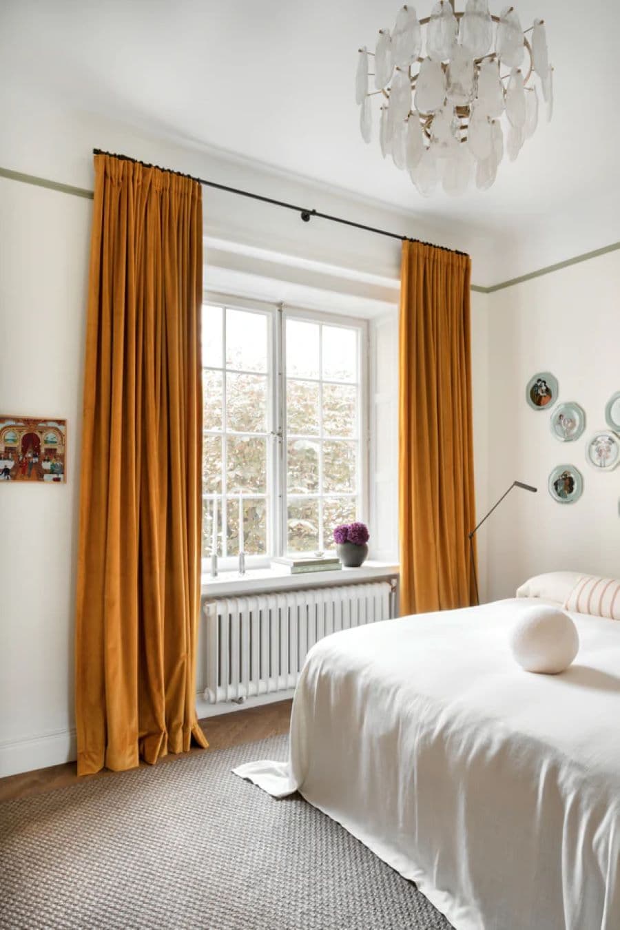 Dormitorio con cortinas de tela de terciopelo en color mostaza, cama con sábanas blancas, lámpara de techo decorativa, y radiador bajo la ventana. Decoración en la pared con platos pintados y una pequeña planta en la repisa de la ventana.