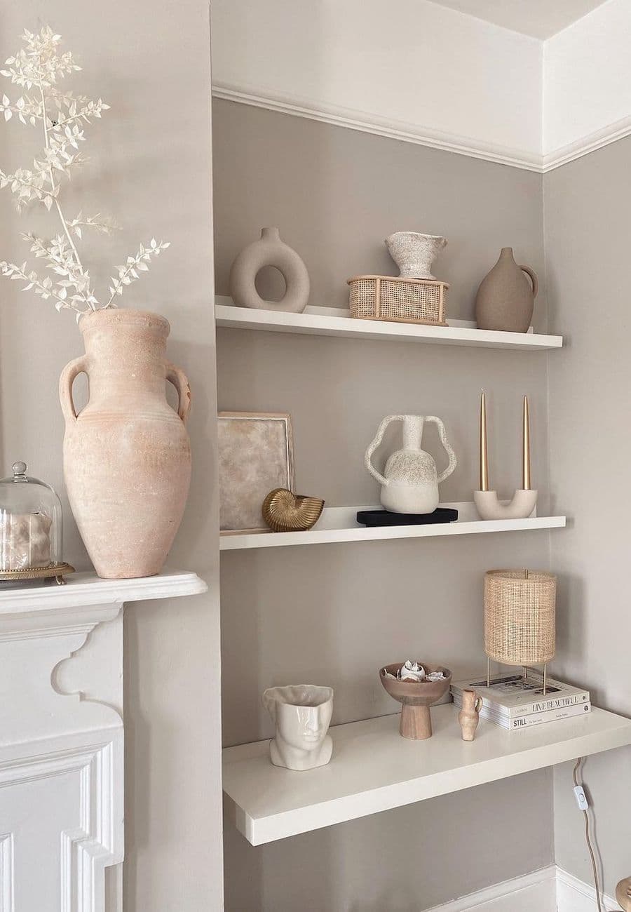 Repisa flotante en un muro color greige. Sobre la repisa hay adornos de cerámica blanca, objetos de fibras naturales, velas y una lámpara. Se ve una chimenea y sobre ella hay un gran jarrón con ramas blancas.
