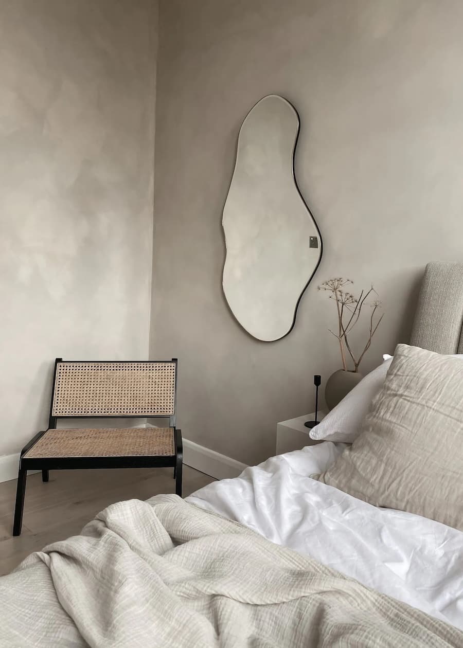 Dormitorio moderno y minimalista con muros de concreto color greige. Cama con textiles blanco y beige, sitial de tejido natural y espejo ondulado en la pared