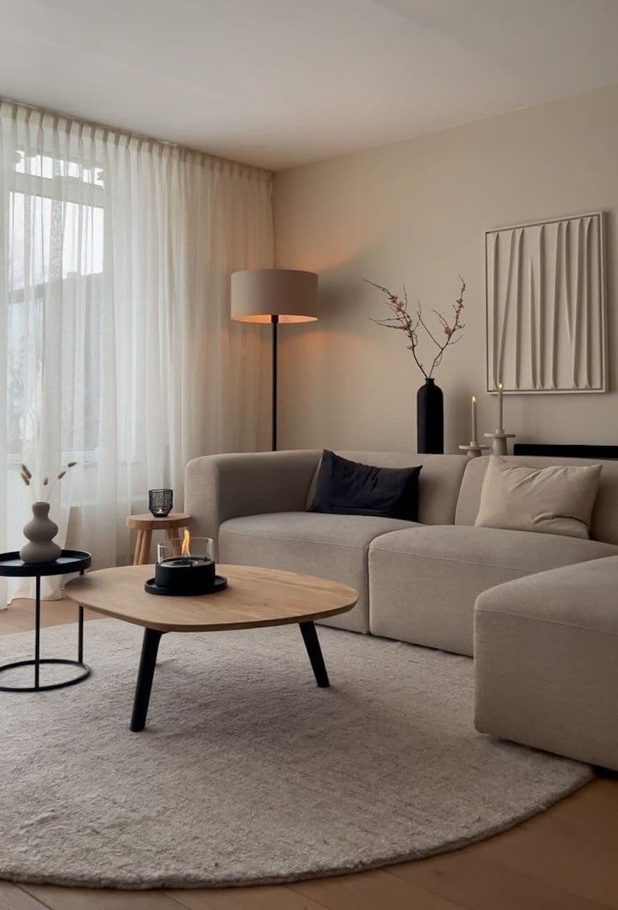 Living con gran sofá en L color greige, con cojines greige y negro. Mesa de centro de madera y pequeña mesa de apoyo negra. Gran alfombra beige. Gran ventanal y lámpara de pie encendida.