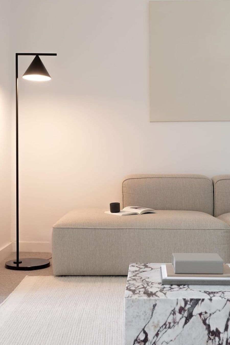 Sala minimalista con sofá de líneas simples, color greige. Mesa de centro y lámpara de pie negra, larga y delgada. Gran cuadro sobre el sofá