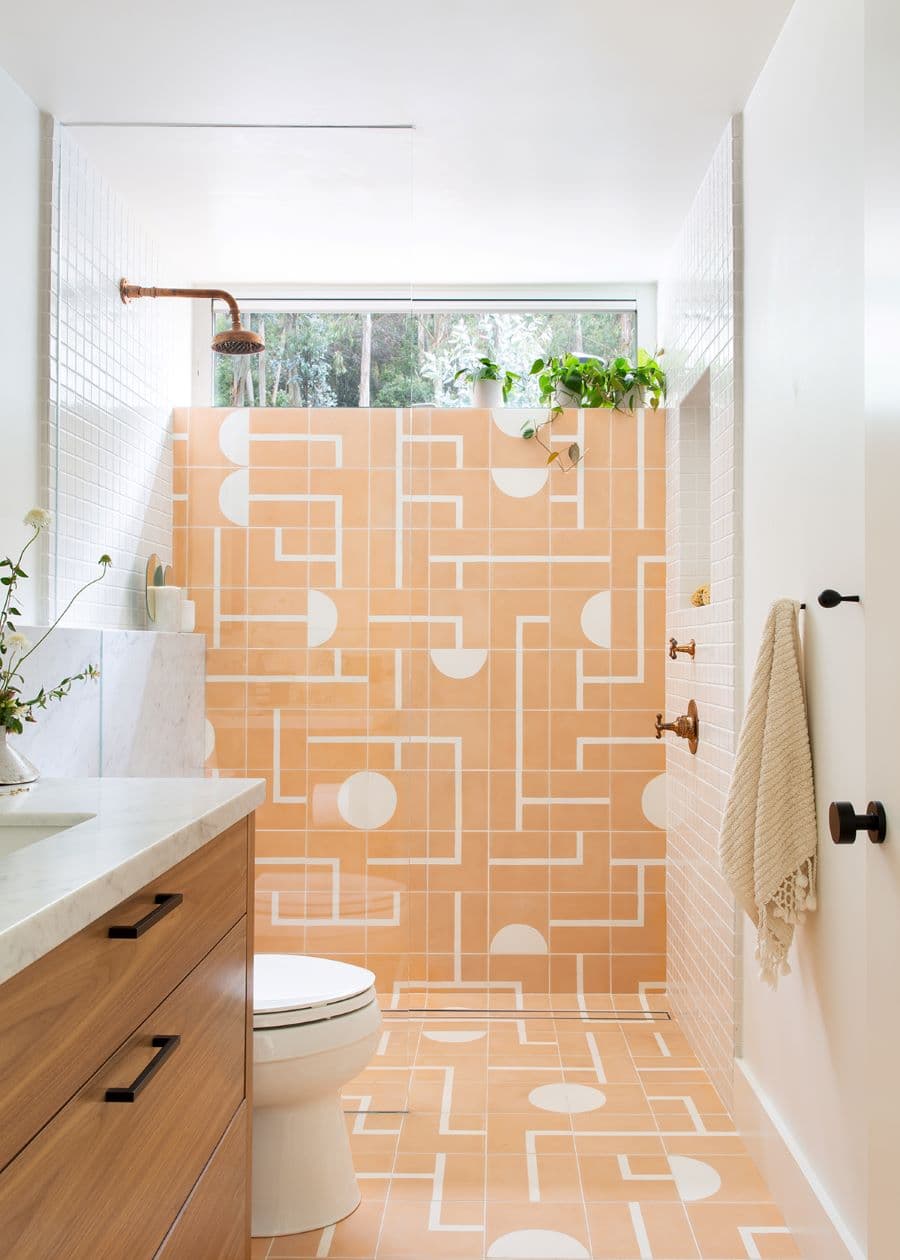 Baño moderno, con azulejos geométricos en color damasco y blanco. También tiene azulejos cuadrados pequeños y un muro pintado blanco. Mampara de vidrio y grifería de cobre. Vanitorio de madera con base de mármol.