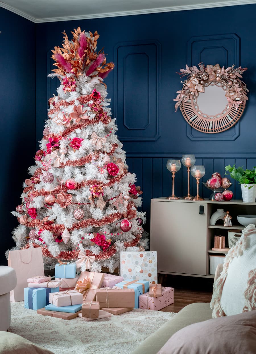 Living con muros azules. Gran árbol de Navidad color blanco con adornos rojo, dorado y rosado. Regalos envueltos en papeles celestes, rosa y rosado.