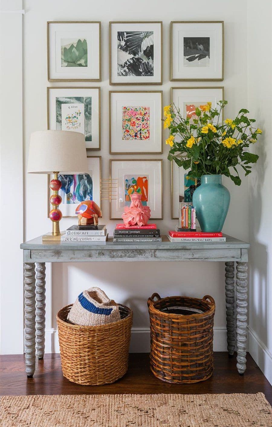 Recibidor de pared blanca con 9 cuadros con ilustraciones de colores, un arrimo celeste pastel, 2 canastos de mimbre, y sobre él, un florero calipso con flores amarillas, una lámpara roja con pantalla blanca, una figura rosada y varias pilas de libros.