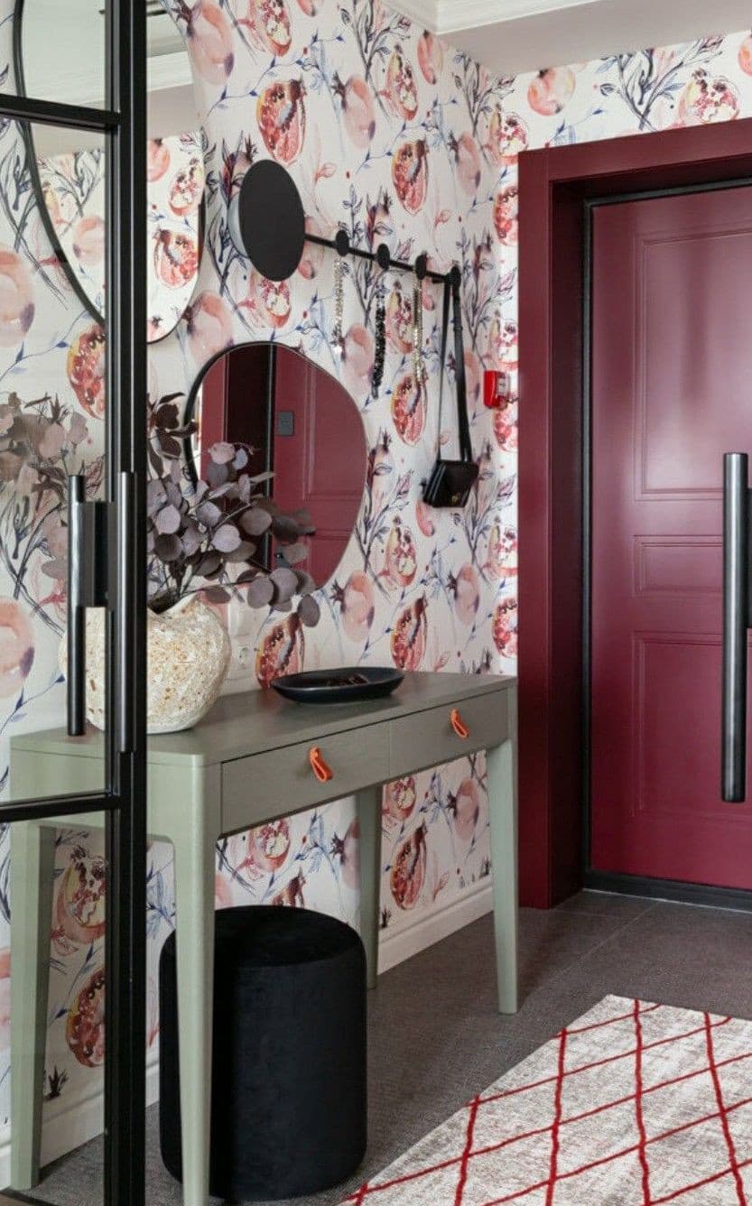 Hall de entrada con un arrimo verde pastel, un espejo sin marco, un florero beige con flores secas, un cuenco negro para las llaves, un perchero negro y papel mural de flores rosadas y grises en el muro.