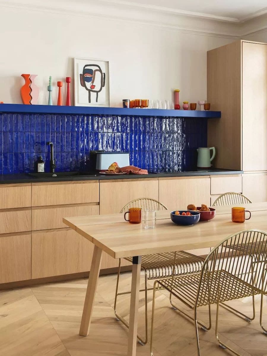 Cocina abierta con muebles de madera clara, azulejos azul eléctrico en la pared y una mesa de comedor de madera clara con sillas metálicas con acabado dorado.