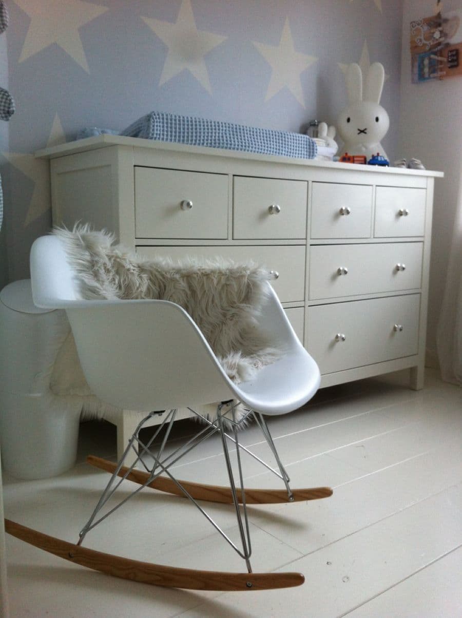 Dormitorio de bebé estilo nórdico. Detalle de silla mecedora eames blanca, con manta peluda sobre ella. Gran cómoda de madera blanca, con cajones