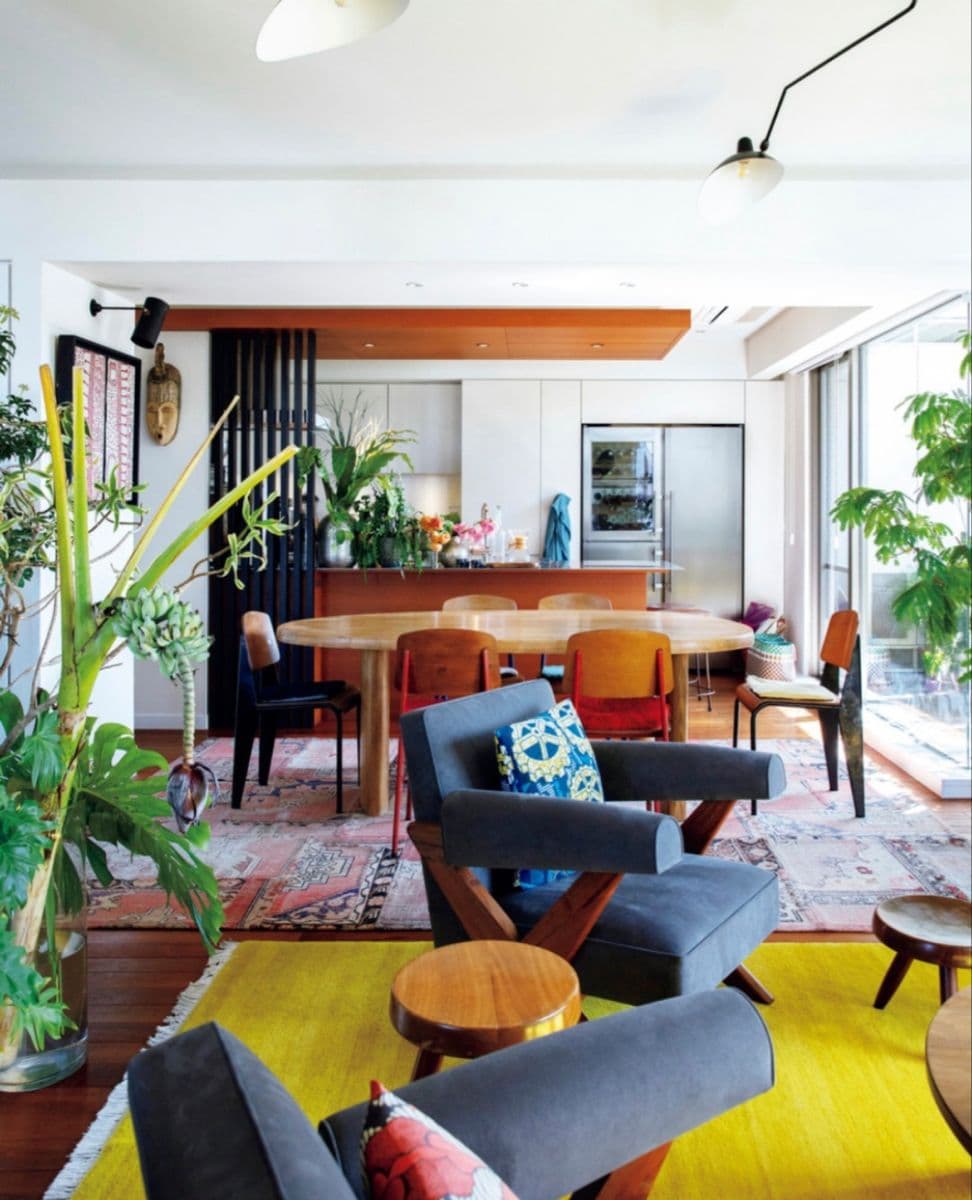 Living comedor estilo vintage moderno. Al fondo hay una cocina y un juego de comedor de madera y, más adelante, se ven poltronas de madera con tapizado color azul marino. Hay una alfombra mostaza, como también una alfombra kilim.