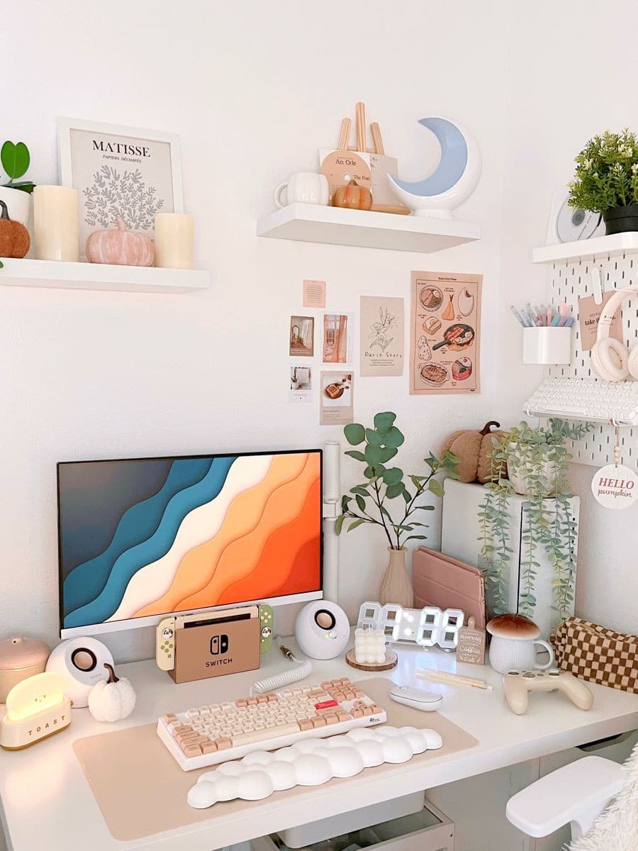 Escritorio blanco con computador, decoraciones, plantas y artículos de escritorio. En el muro hay repisas flotantes blancas