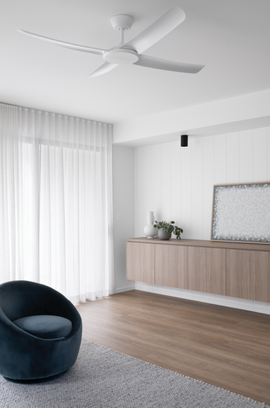 Living estilo moderno y minimalista. Muros blancos y gran ventanal con visillo. Mueble flotante de madera, alargado. Sitial redondo, de terciopelo azul. Ventilador de techo blanco de estilo contemporáneo.