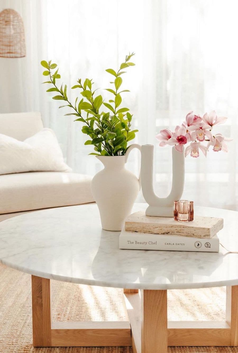 Mesa de centro redonda, de mármol con un florero blanco con ramas verdes, libros y un florero en forma de U con flores rosadas.