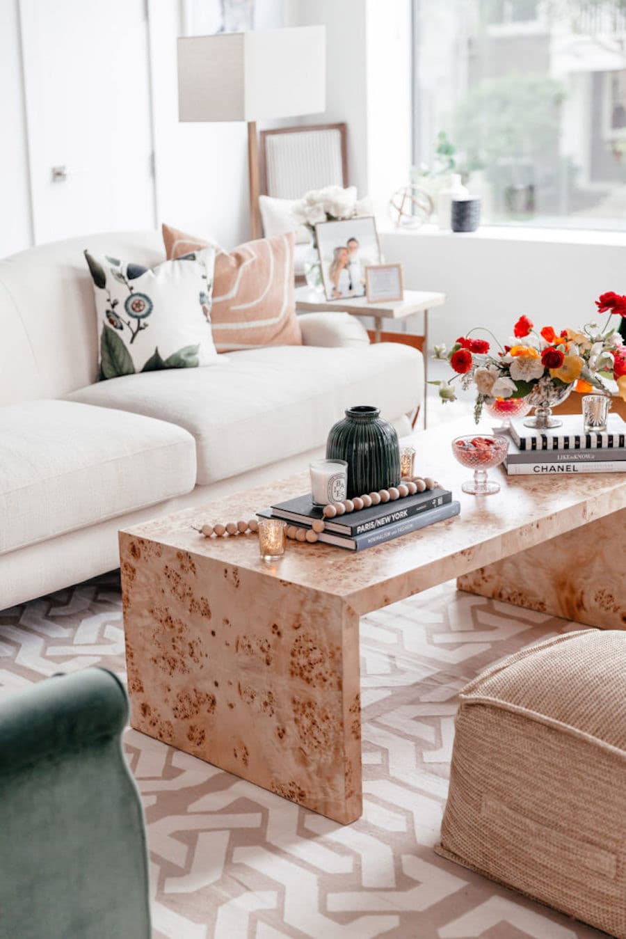 Mesa de centro de mármol rosa con libros, adornos, un jarrón negro y ramo de flores de colores. Sofá blanco y pouf rosa