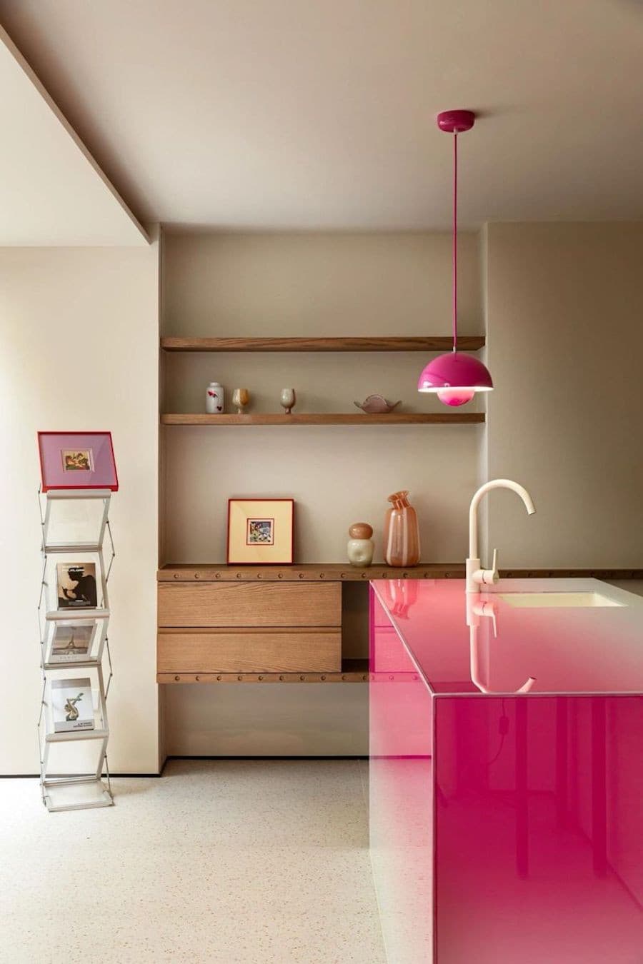 Departamento moderno de paredes beige, concepto abierto, un mueble de madera anclado a la pared como estante y una isla de cocina de color fucsia. Sobre ella hay una lámpara colgante del mismo color.