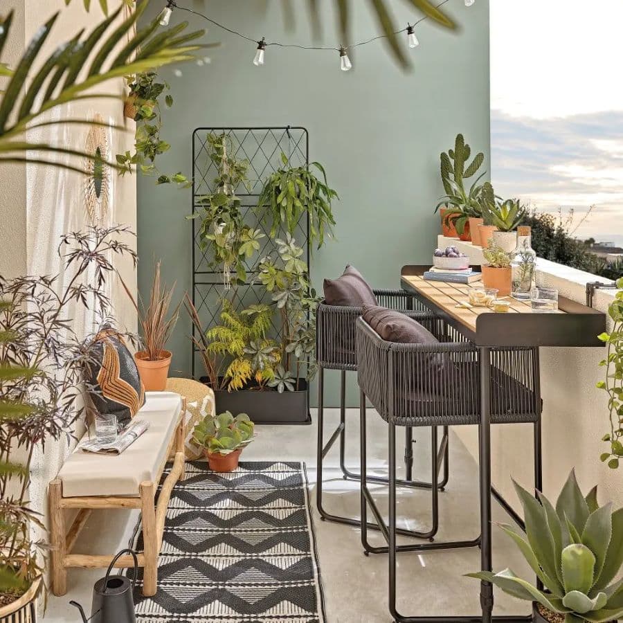 Terraza con barra en la baranda, de madera con metal. Dos sillas altas de metal negro, con respaldo y cojines. Banqueta de madera y cojín blanco, pegada al muro. La terraza tiene un muro verde y otros blancos. Hay una alfombra negra, gris, y blanca