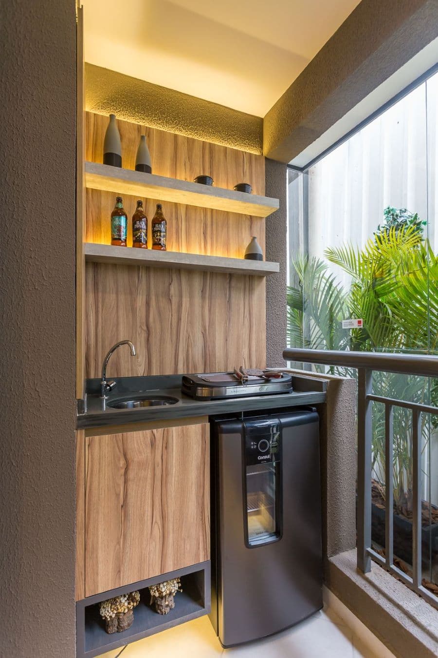 Rincón de terraza con un pequeño mueble de cocina de madera, que tiene lavaplatos, parrilla y en la parte de abajo, un mini bar o frigobar. Arriba tiene dos repisas flotantes con luces. La terraza tiene banda negra y está cerrada con vidrios.