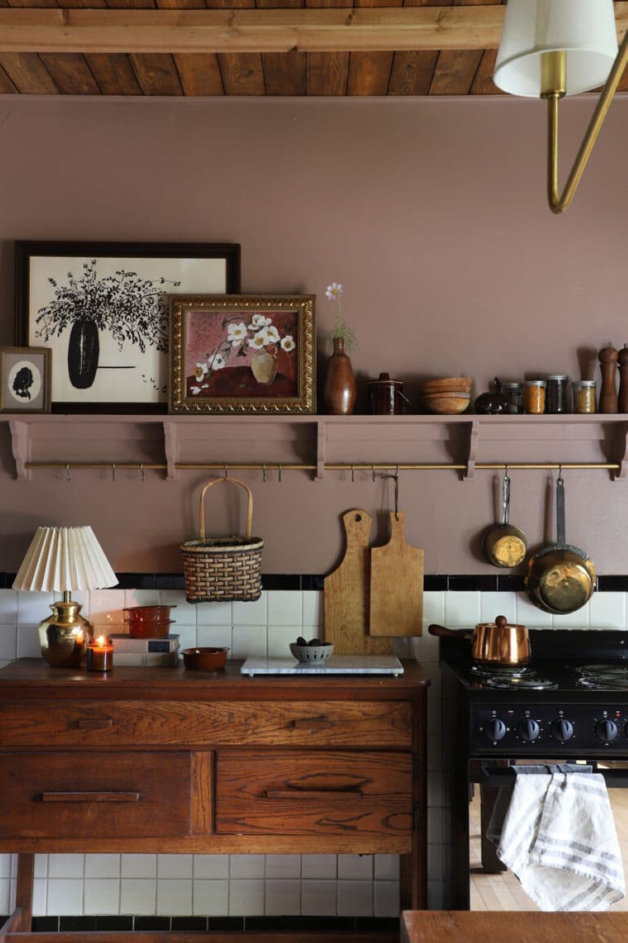 Cocina rústica con muro pinta de color café rojizo. Gran repisa de madera flotante con vasijas de cerámica y cuadros de flores. Muebles de madera vintage, tablas de madera, ollas de cobre y una pequeña lámpara