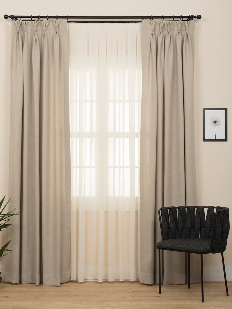 Ventana con cortinas dobles. Visillo beige translúcido y cortinas de tela beige. Al costado hay una silla negra y una planta.