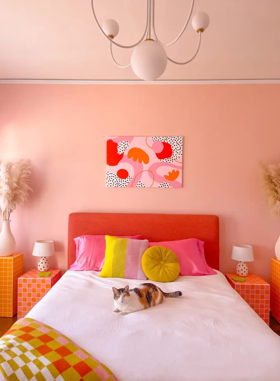Dormitorio estilo dopamina. Muros color durazno. La cama está al centro con un plumón blanco, una piecera rosa y almohadas y cojines color fucsia, rosado y amarillo. El respaldo de la cama es naranjo. Hay dos veladores naranjos con lámparas pequeñas