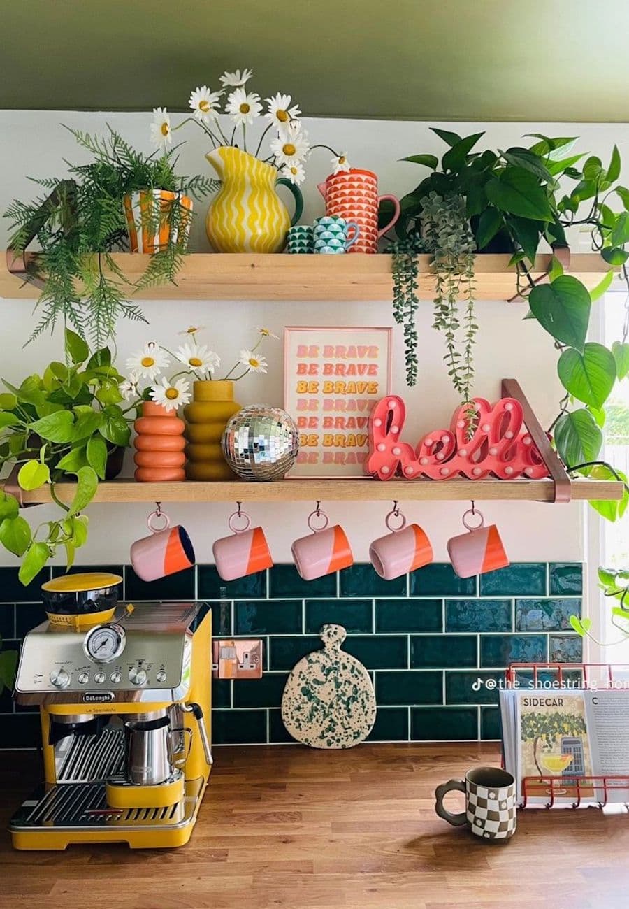 Cocina estilo dopamina. Muro en la parte inferior con cerámicos estilo subway color petróleo. Arriba, muro blanco con dos repisas flotantes de madera con adornos de colores vibrantes, cuadros y plantas. Tazones colgando y máquina de café amarilla