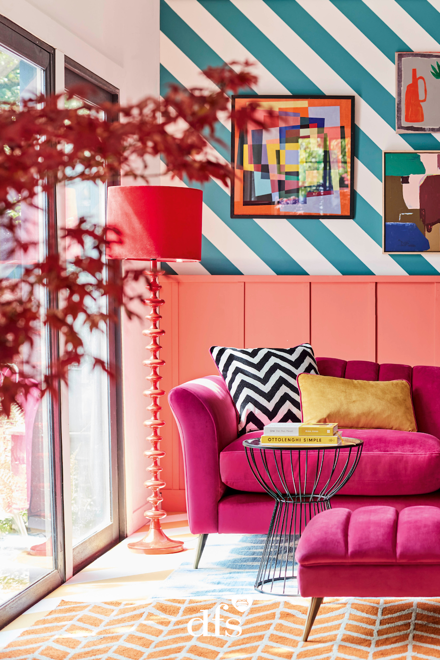 Living estilo dopamina. Muro en la parte baja con revestimiento de madera color salmón. En la parte superior papel mural rayado, azul y blanco. Sofá fucsia con cojín zig zag blanco y negro y amarillo. Mesa de apoyo redonda, negra, de metal. Lámpara de pie 