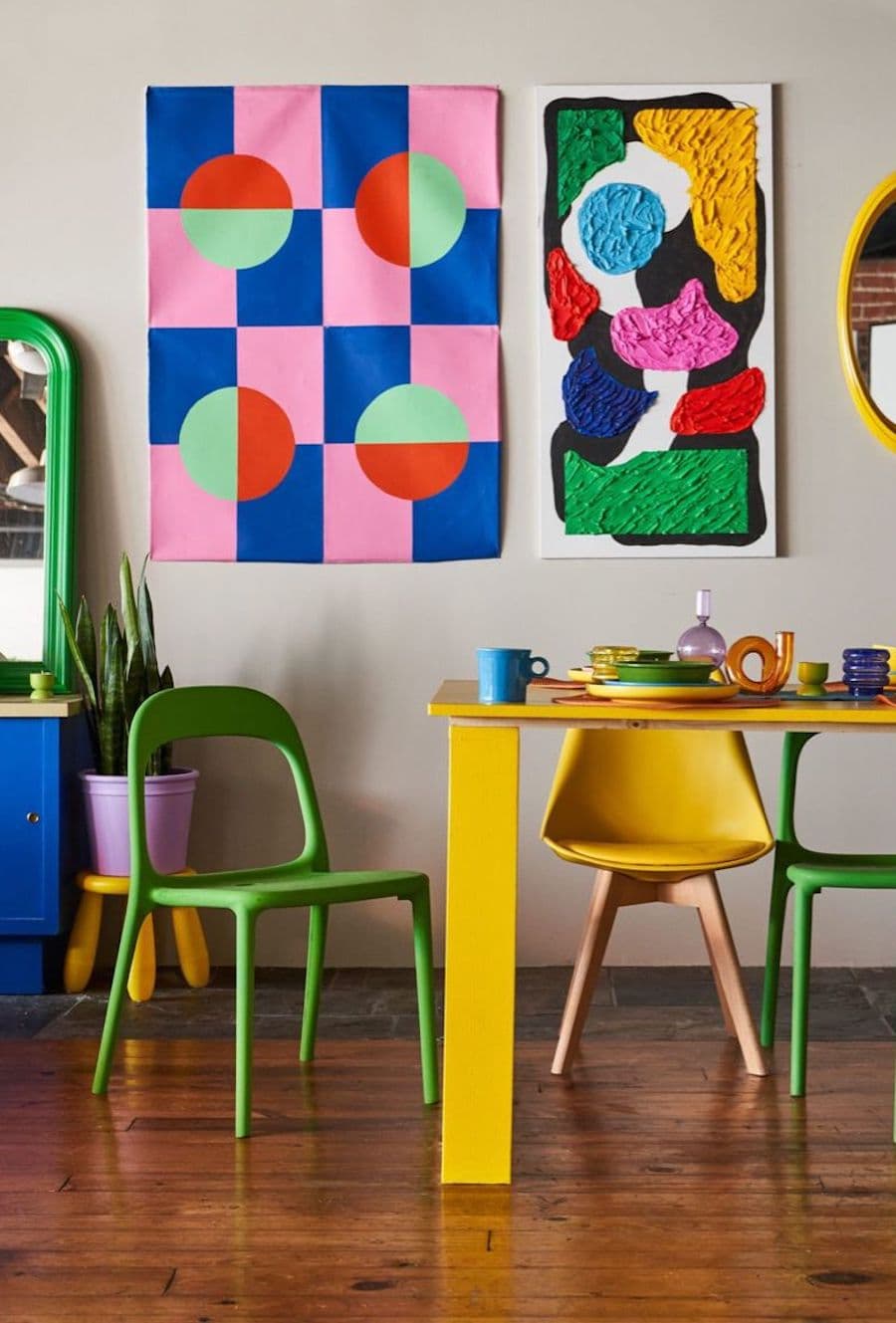 Comedor moderno, estilo dopamina. Mesa amarilla con dos sillas verdes y una estilo eames color amarillo. En el muro gris claro hay dos imágenes grandes con dibujos de colores abstractos y geométricos. El piso es de madera oscura