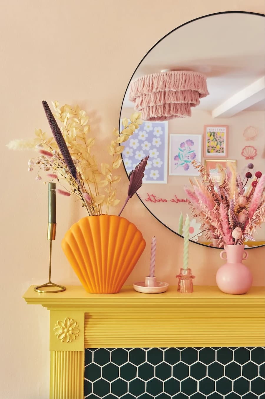 Chimenea empotrada con marco de color amarillo. Sobre ella hay portavelas rosados de diferentes tamaños y uno dorado. También hay floreros, uno grande de color naranjo y otro tipo vasija color rosado, ambos con flores secas. Espejo redondo, muro damasco