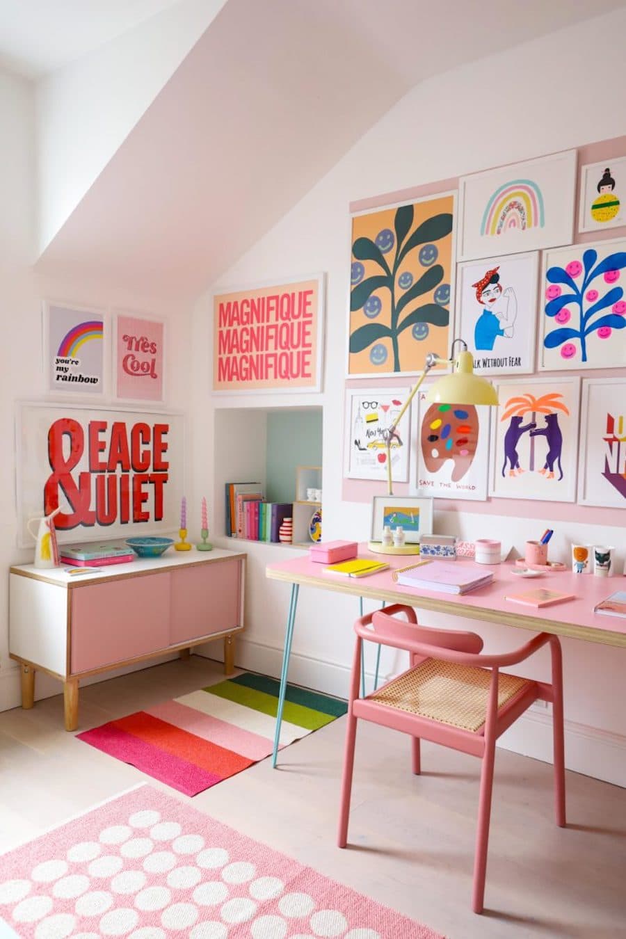 Escritorio estilo dopamina. Muros blancos, mesa rosada con patas celeste, silla rosa. Muro galería con cuadros de colores y estilo moderno. Arrimo con puertas rosadas, pequeña alfombra con líneas de colores.