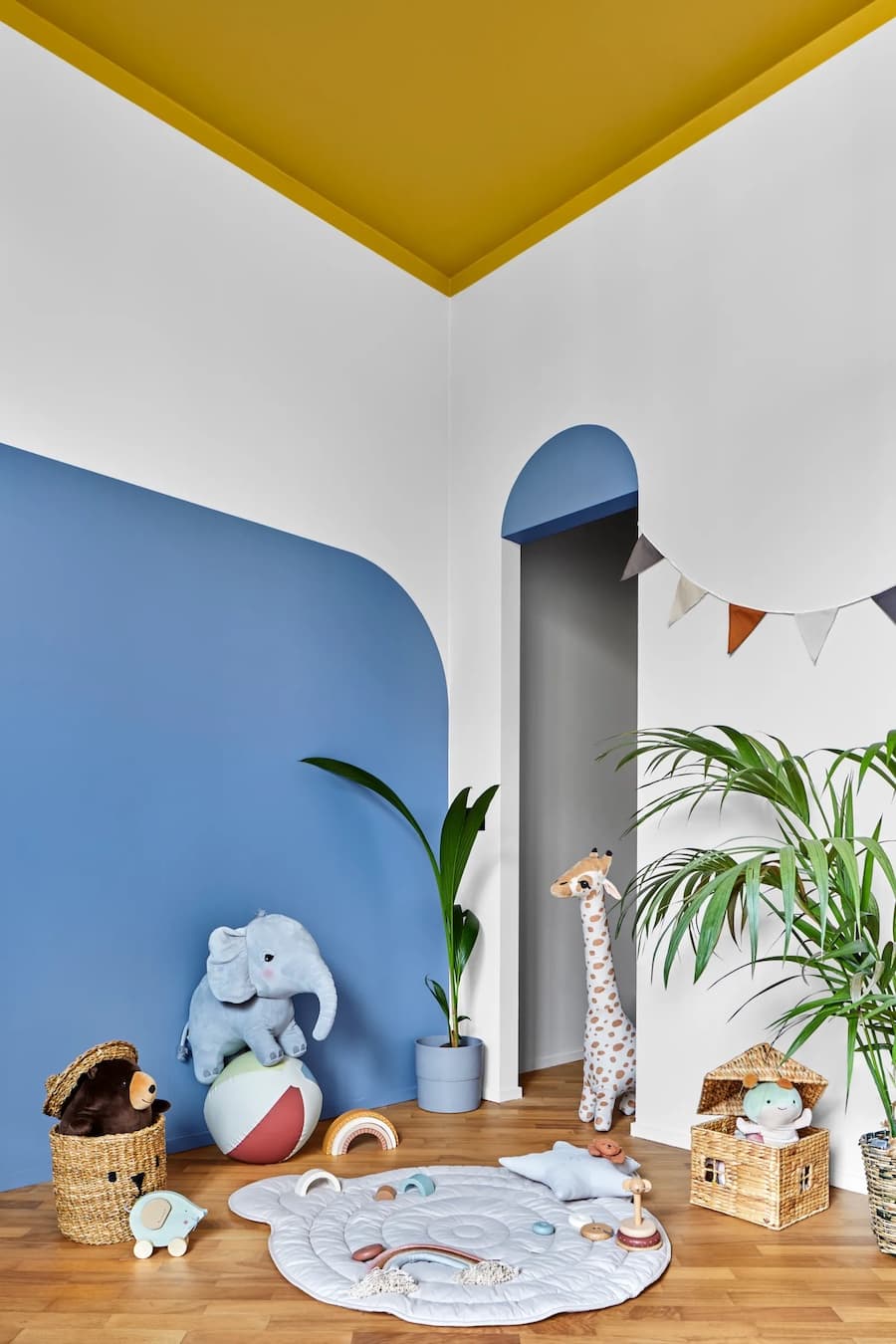 Dormitorio infantil con paredes blancas y un muro pintado azul con forma curva. El techo es color ocre y traspasa los muros para darle continuidad. Juguetes y animales de peluche en el suelo, y plantas tropicales en macetas de colores