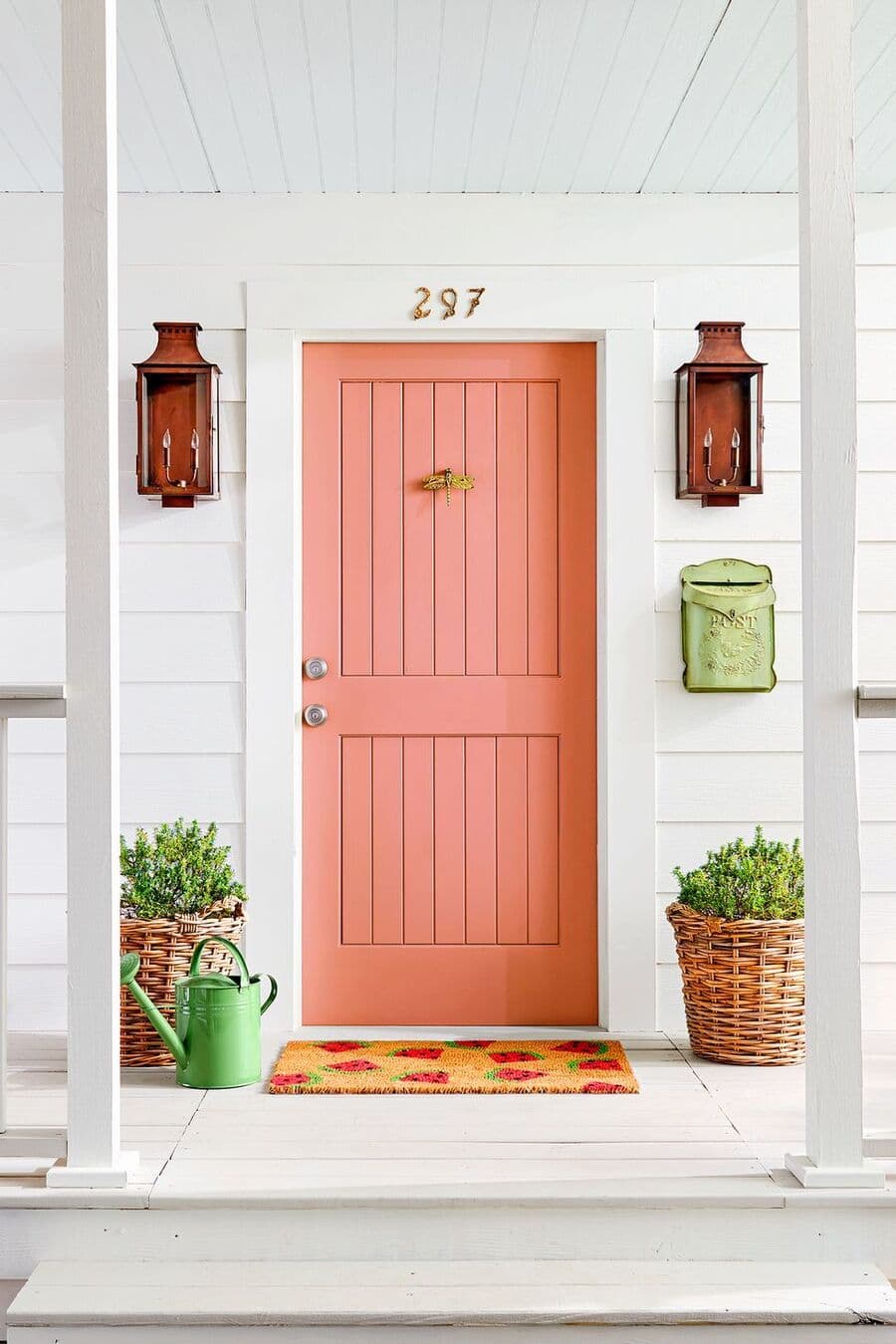 Entrada de una casa con una puerta de madera color sandía. Dos focos de vidrio color ámbar a cada lado, un pequeño buzón verde y dos macetas de mimbre con plantas, Una regadera verde en el suelo junto al limpiapiés