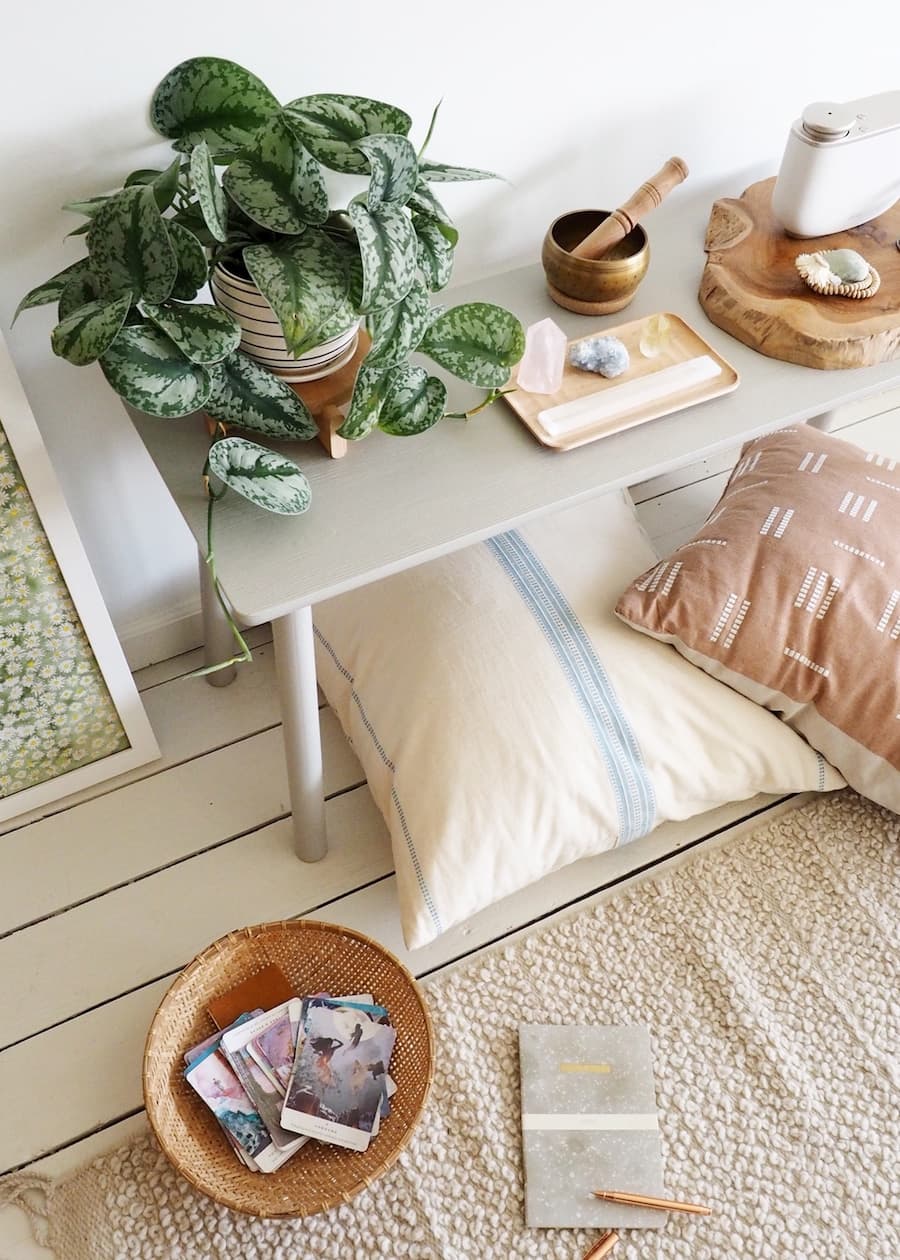 Rincón zen con una banqueta blanca. Sobre ella hay plantas y un pequeño altar con piedras y objetos para meditar. Bajo la banqueta hay cojines un pequeño canasto de mimbre y una alfombra beige.