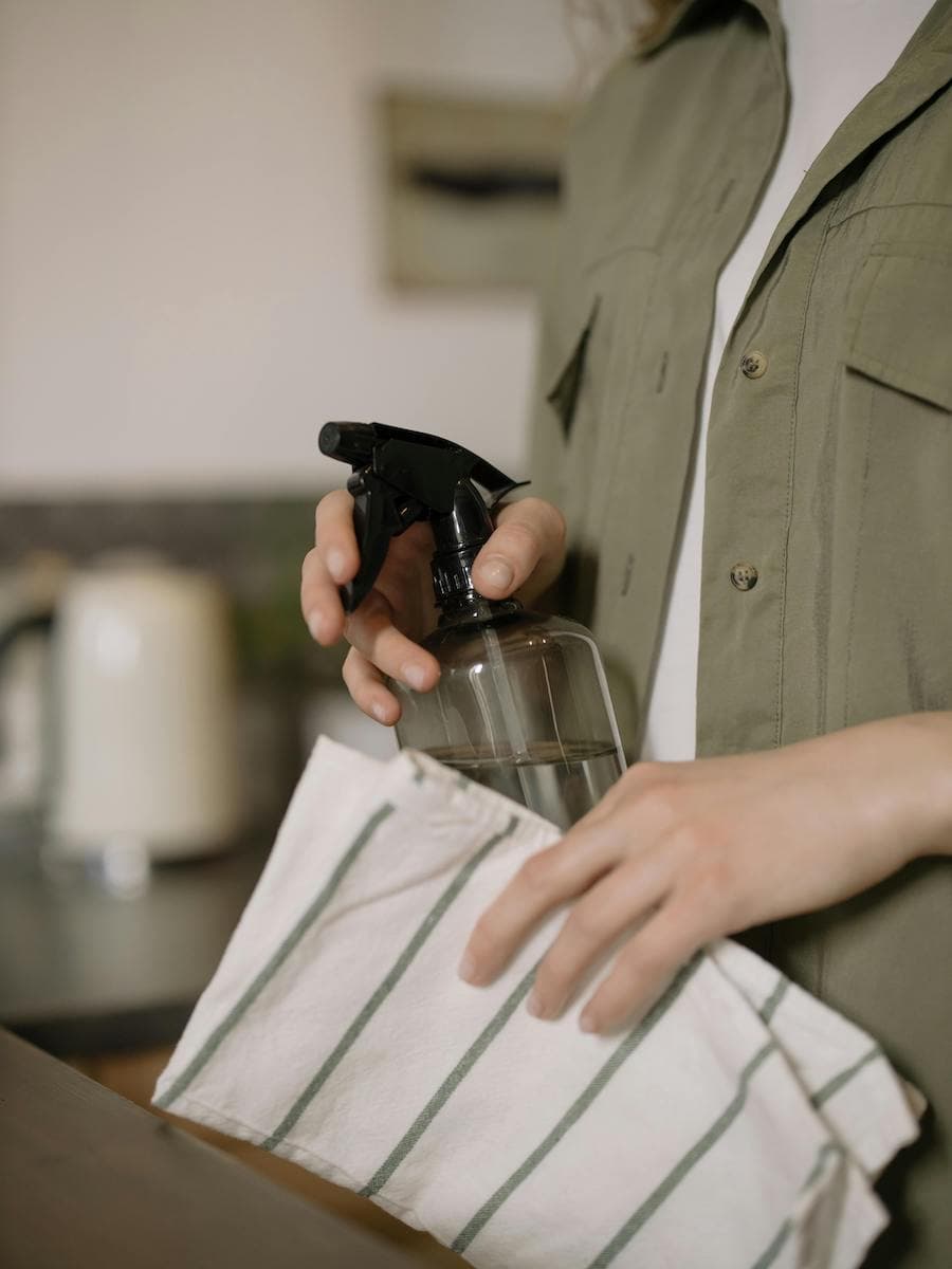 Tips de limpieza, detalle de una persona con un bolso de tela beige y en la otra mano un spray con una botella trasparente.