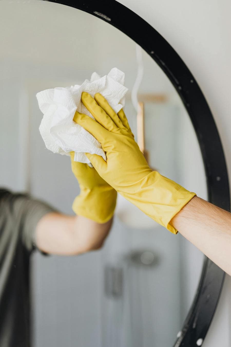 Tips de limpieza, detalle de una mano con un guante amarillo limpiando un espejo redondo con un paño blanco.