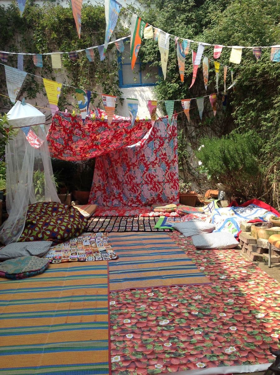 Carpa hecha con telas en un patio. Al lado hay un mosquitero blanco y del techo cuelgan guirnaldas de banderines de colores. En el suelo hay 4 alfombras grandes de diferentes colores y diseños. También hay cojines de diferentes tamaños y formas.