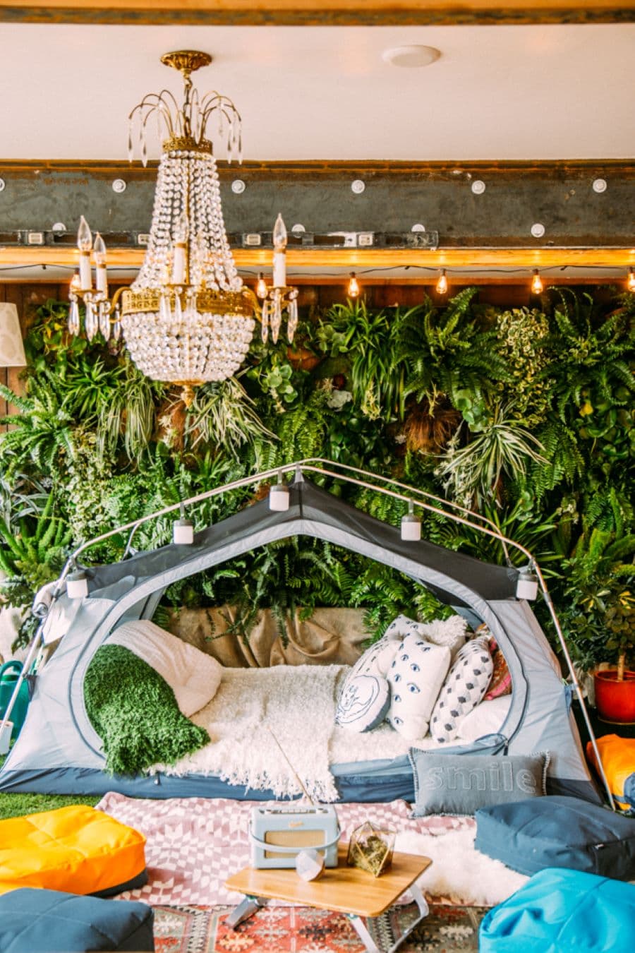 Carpa dentro de una casa. Está abierta y dentro tiene colchones y cojines. Afuera hay alfombras en el suelo. En la parte de atrás hay un muro verde con plantas y del techo cuelga una lámpara de lágrimas de cristal.