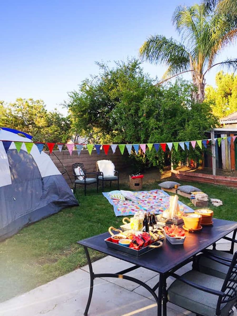 Gran patio con una carpa armada. Mesa de jardín negra con comida y bebidas. Sobre el pasto hay una manta floreada y dos sillas negras. Una guirnalda de banderines cruza el espacio