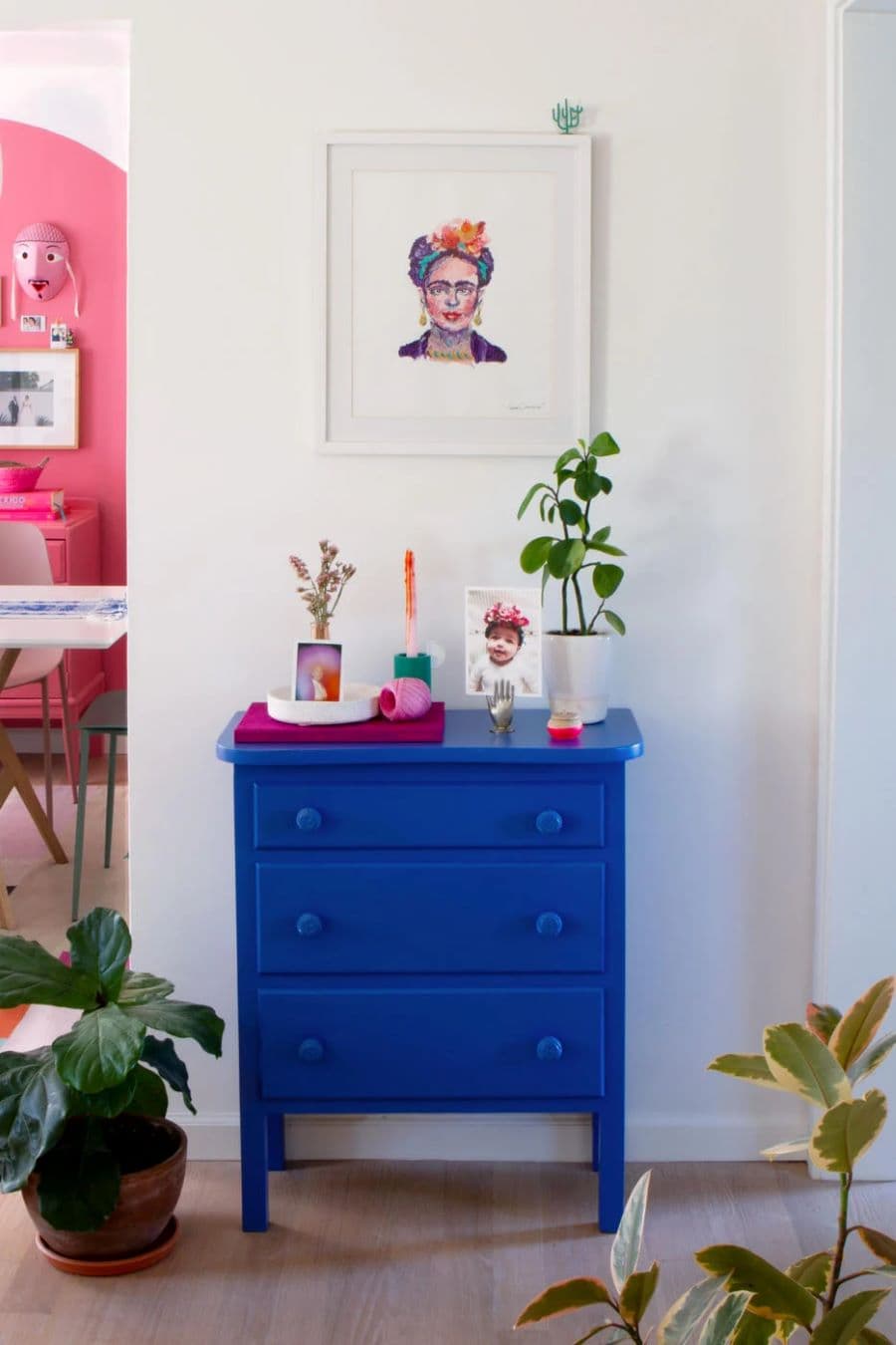 Hall de entrada con una pequeña cajonera con tres cajones de color azul cobalto. Sobre ella hay adornos, fotografías y una planta. En el muro blanco hay un cuadro con una imagen de Frida Khalo. A los costados hay plantas