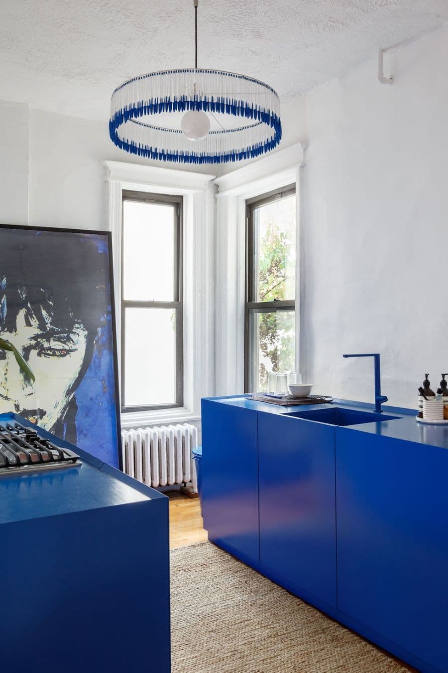 Cocina de estilo moderno en un departamento antiguo. Muebles azul cobalto. Gran cuadro apoyado en la pared con el retrato de un hombre. Lámpara de techo redonda, hecha con lápices con tapa azul y gran ampolleta blanca. Dos ventanas amplias sin cortinas