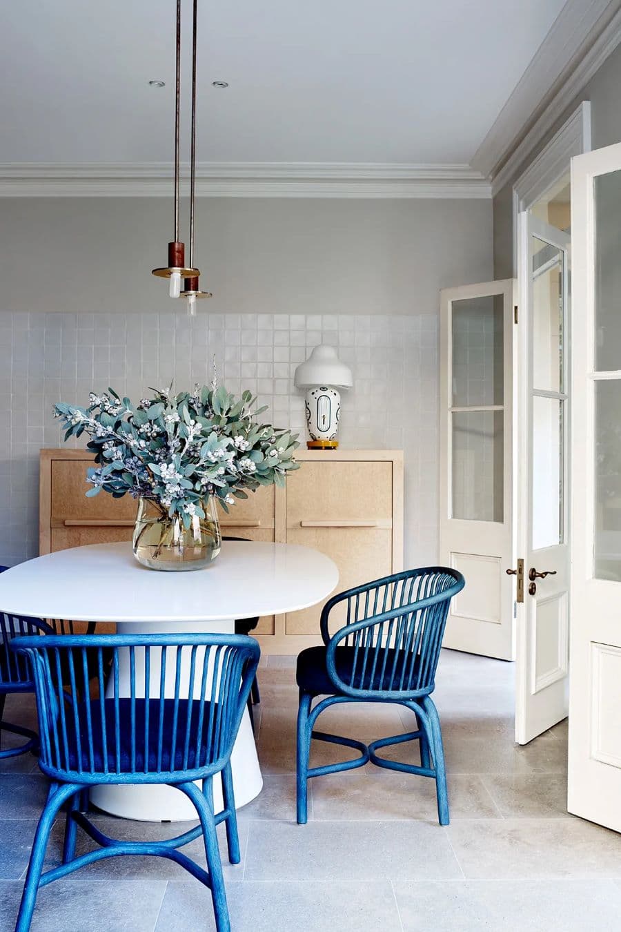 comedor moderno con mesa redonda blanca y dos sillas de ratán azul cobalto. Gran ramo de flores sobre la mesa en un jarrón de vidrio. Atrás hay un mueble con puertas de madera. Lámpara de techo doble dorada con ampolletas a la vista