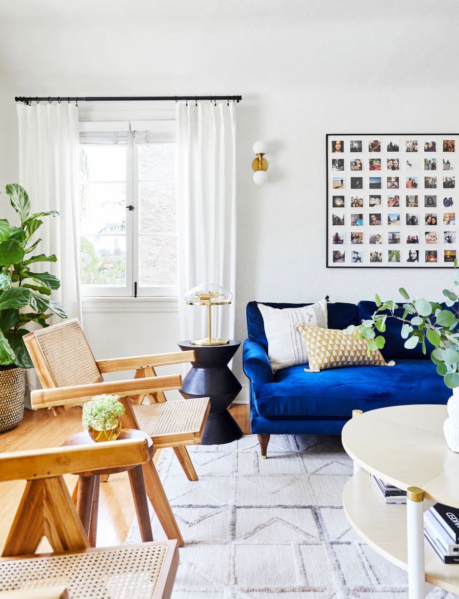 Living de estilo clásico moderno. Sillón de terciopelo azul cobalto, con dos cojines. Dos sitiales de madera y respaldo de cannage. Gran alfombra beige con líneas. Junto al sofá hay una mesa de apoyo negra con formas cónicas. Gran cuadro con fotos