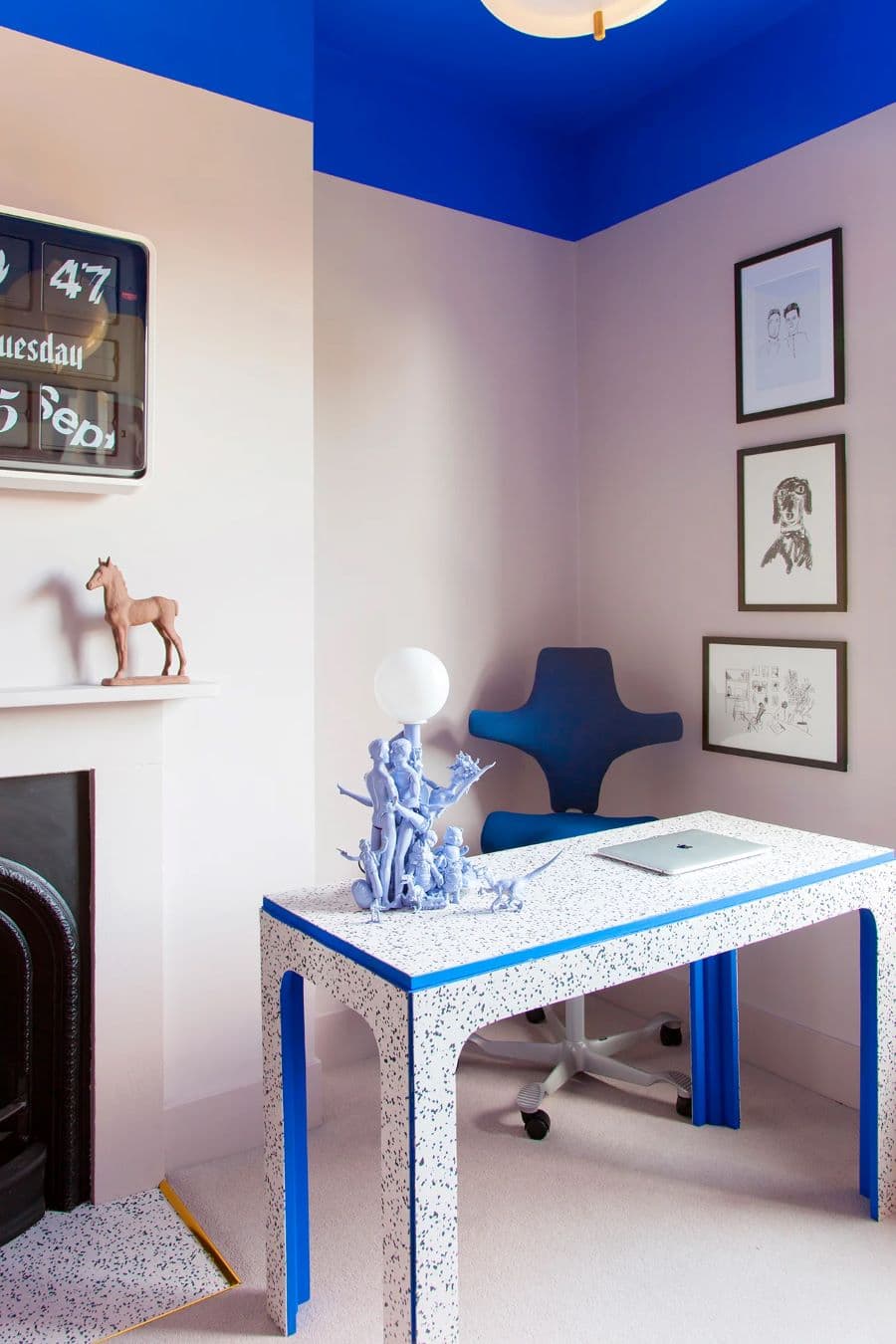 Mesa de escritorio tipo terrazo, de base blanca y líneas azules en el contorno. Silla de escritorio modera, azul cobalto. Los muros son rosa pálido y el techo está pintado de azul cobalto. En el muro hay tres cuadros de marco negro, con ilustraciones.