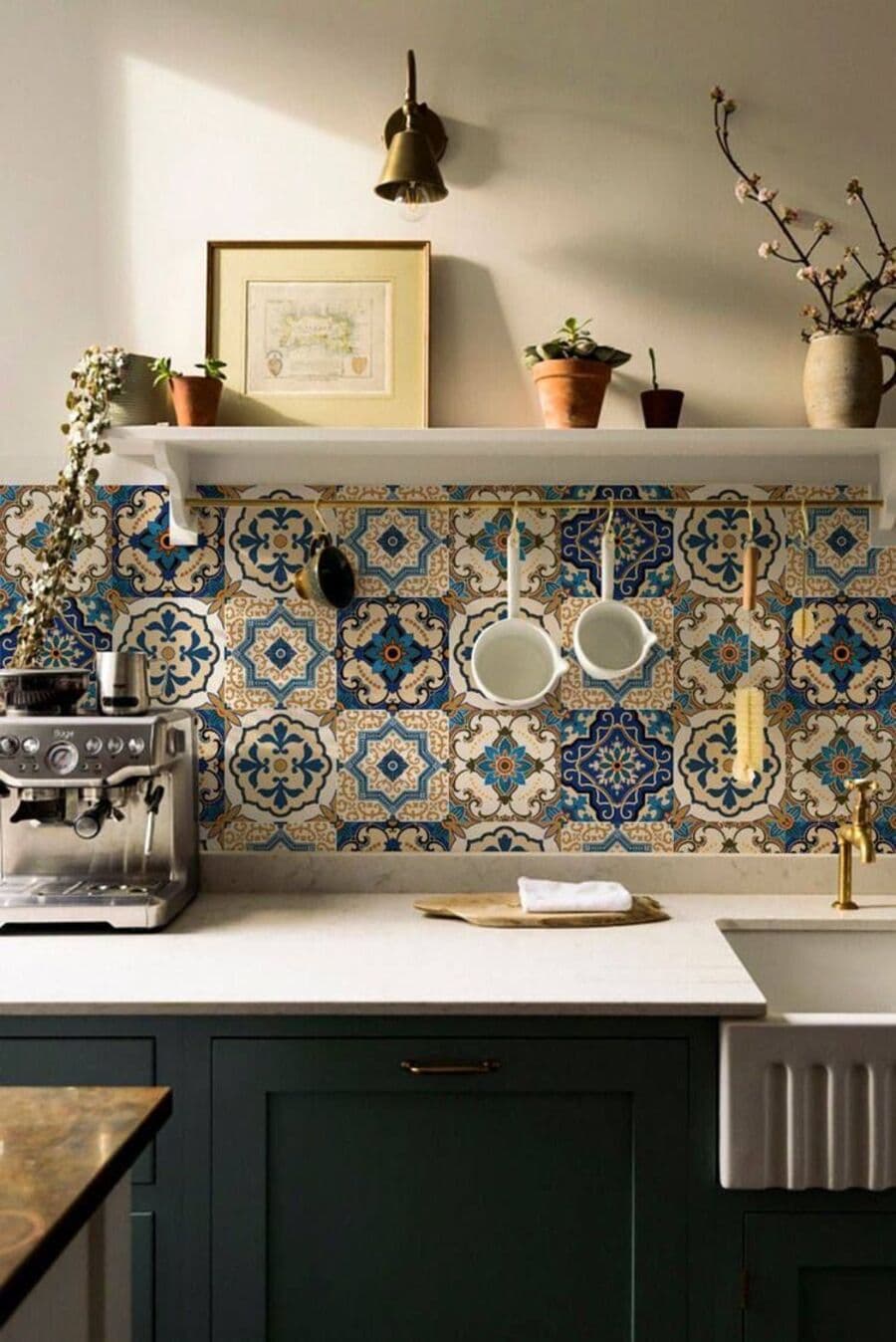 Detalle de cocina estilo tradicional, con baldosas hidráulicas en el muro. Son de color azul y beige. Muebles de cocina color negro y mesón blanco. Sobre las baldosas hay una repisa flotante blanca con un cuadro y plantas. Cafeteracromada